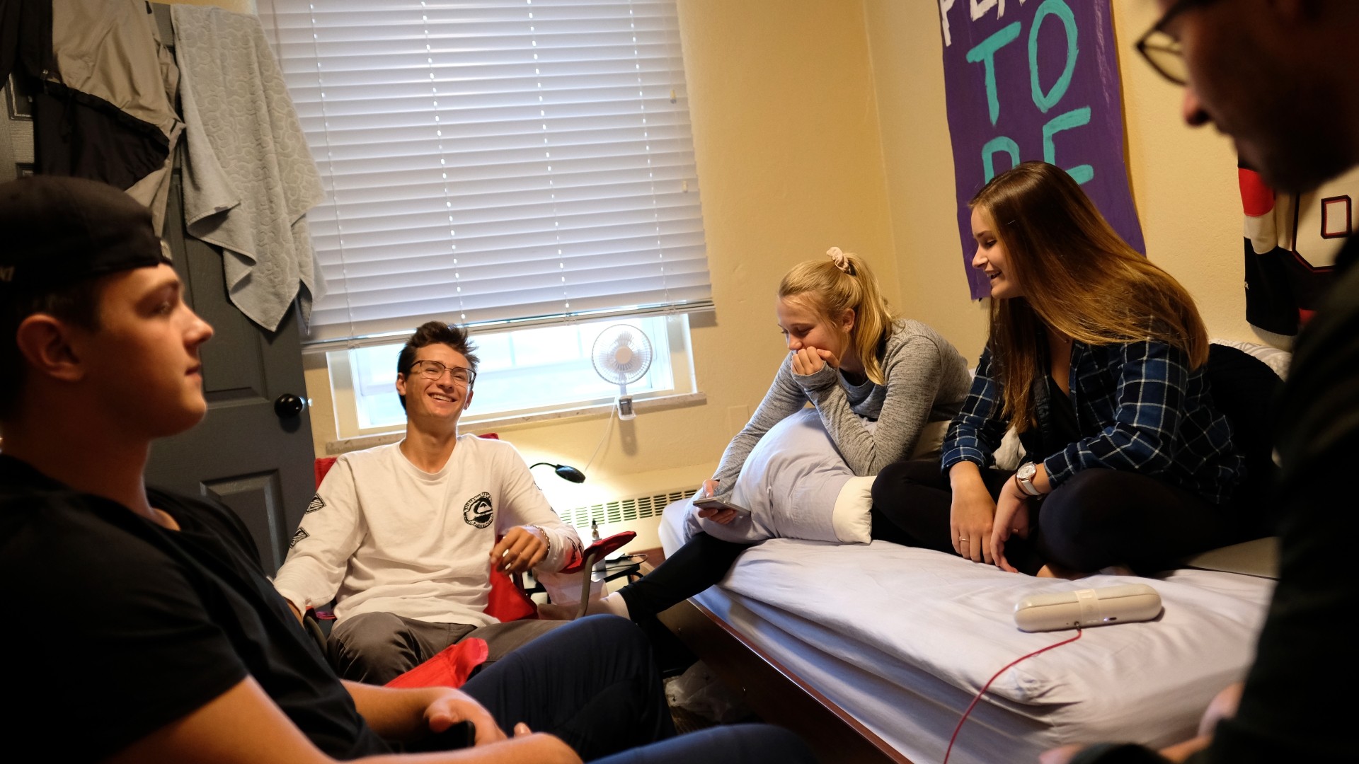 Students socializing in a residence room. Two students are sitting on the bed, while the others are sitting on chairs.