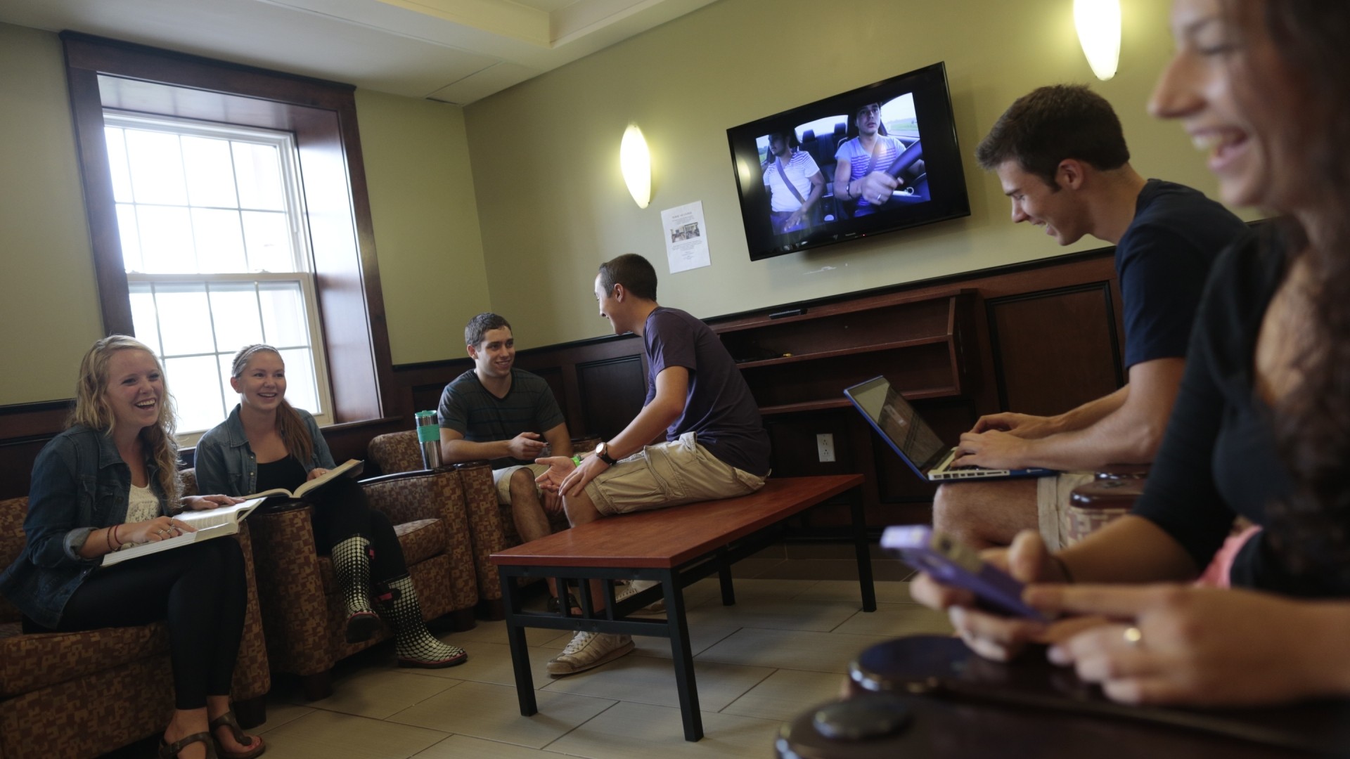 Students interacting in the Bishops Hall lounge area. There are multiple sofa chairs, a coffee table and a TV on the wall.