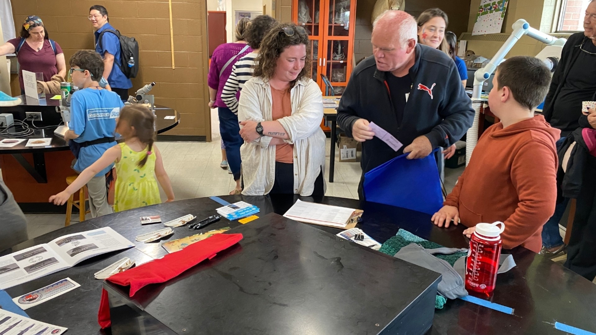 People engaged in various activities around tables in a classroom or workshop with shelves and cabinets in the background.