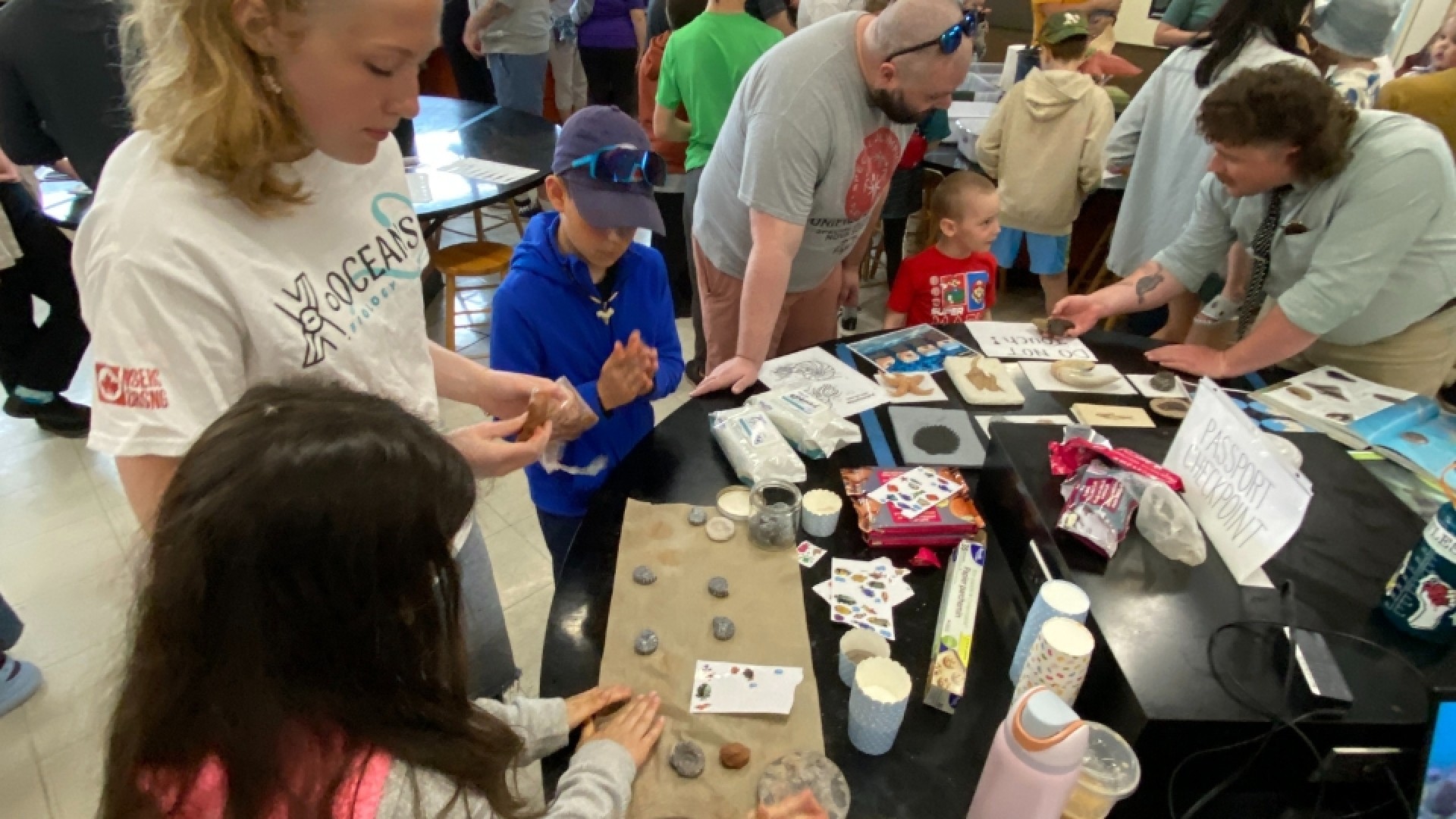 People gathered around a table with fossils, pamphlets, and cups at an indoor educational event.