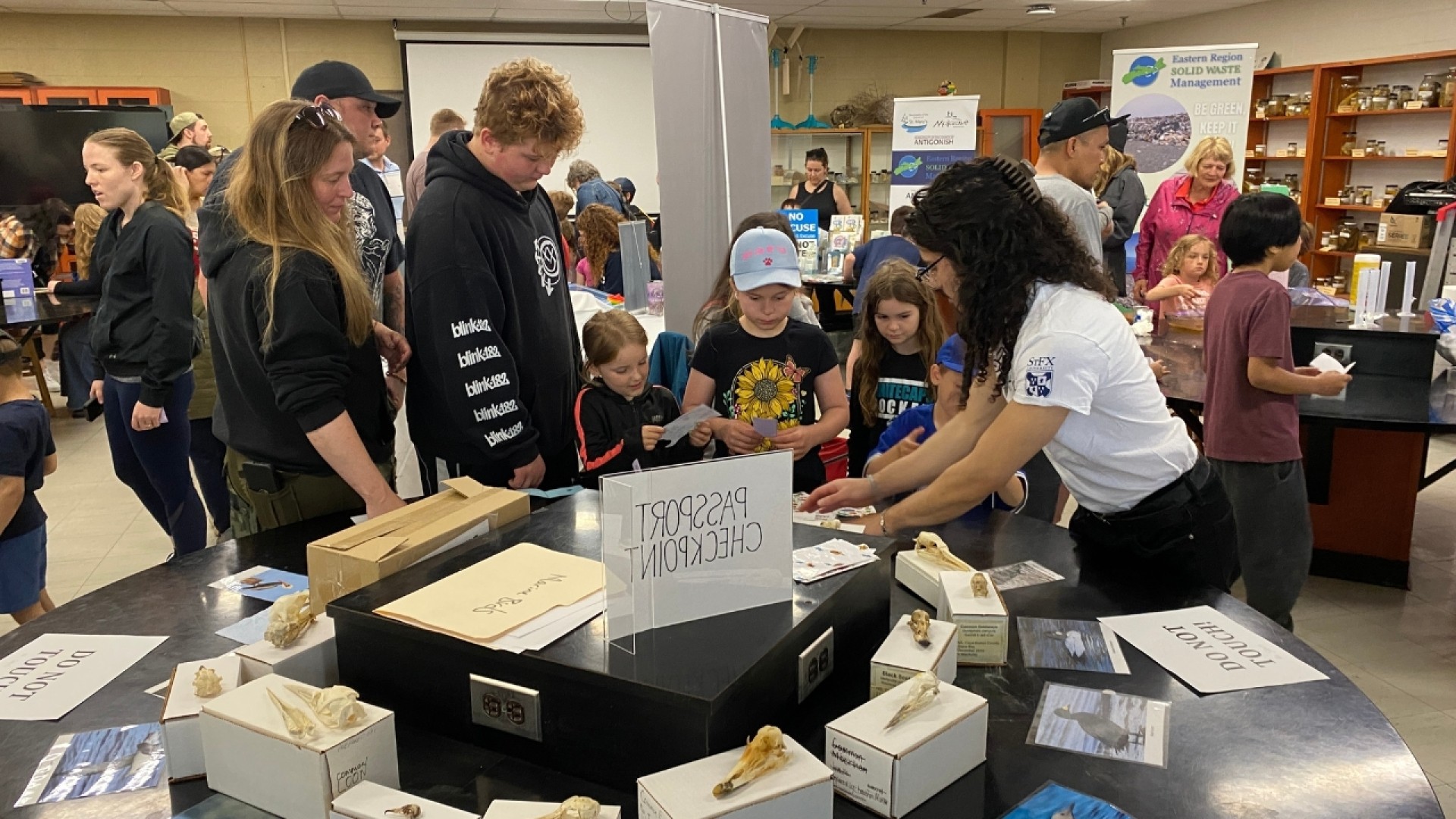 Children and adults gathered around a table with display boxes and a 'PASSPORT CHECKPOINT' sign at an educational event.