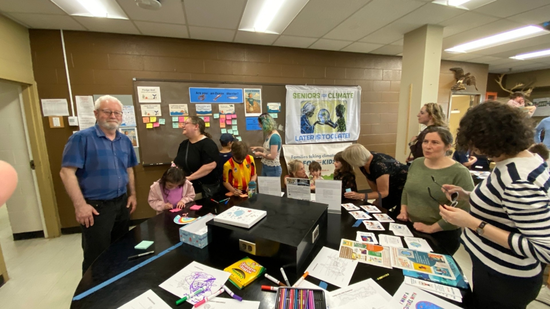People in a classroom working on art activities at a table with a 'Seniors 4 Climate' banner and posters in the background.